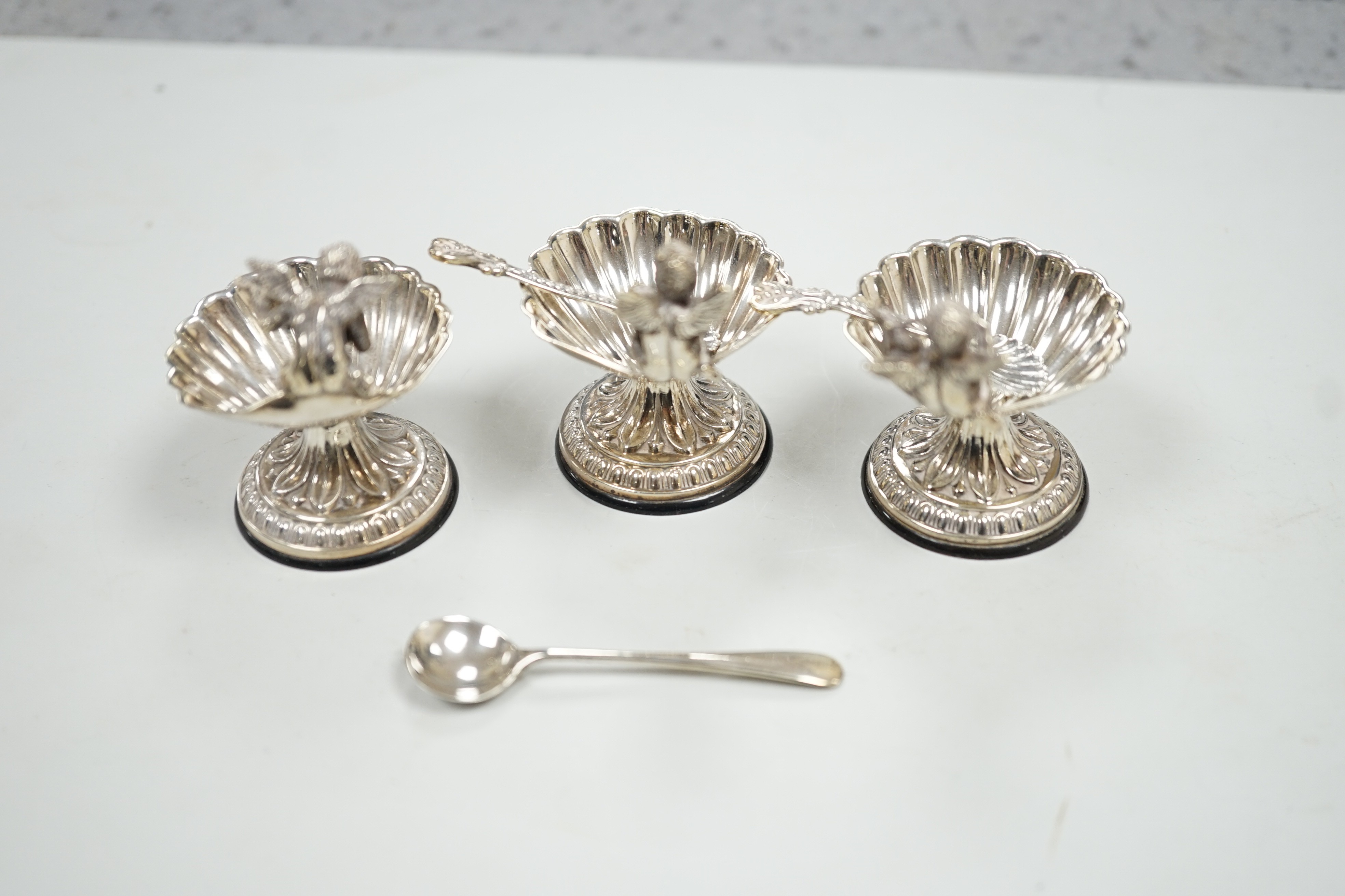 A set of three 20th century Spanish white metal shell pedestal salts with cherub handles and three spoons, height 50mm.